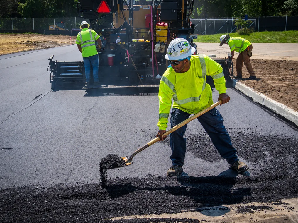 Crew member shoveling hot mix asphalt in front of a paver on a commercial lot, parking lot paving