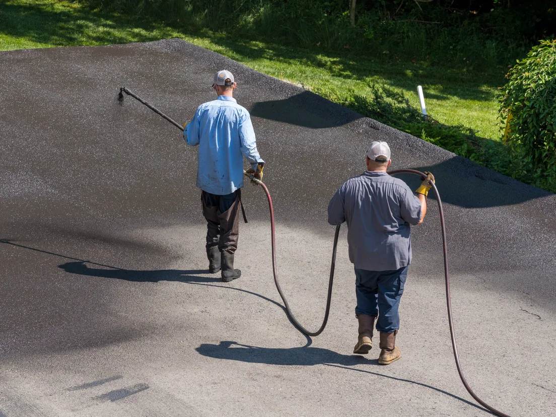 Two workers spraying sealcoat onto a residential asphalt driveway, sealcoating
