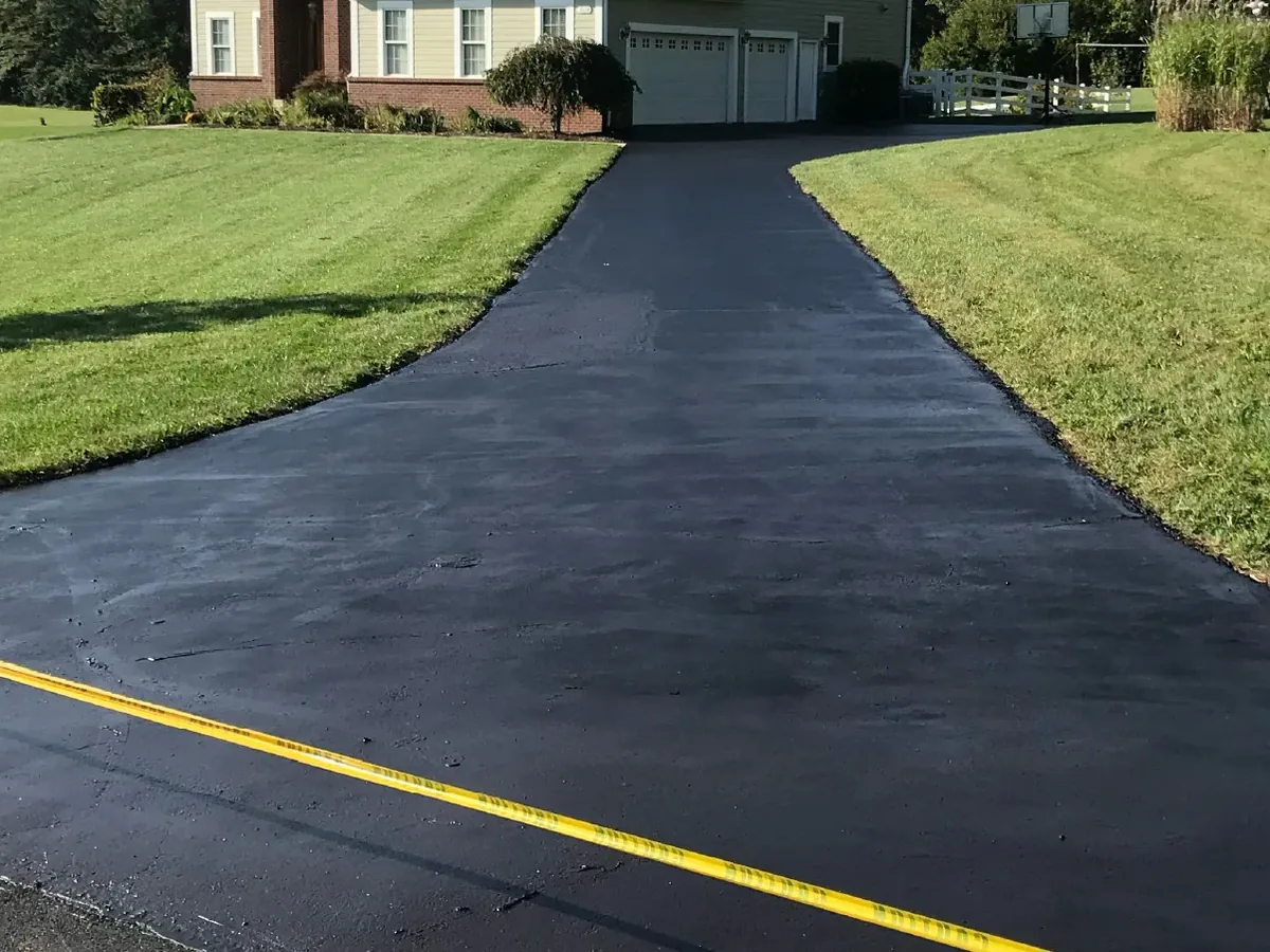 Freshly sealcoated asphalt driveway between lawns with caution tape across the end, sealcoating